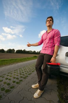 Young man with black reserve canister on a lonely country road leans helplessly against a car and waits for help