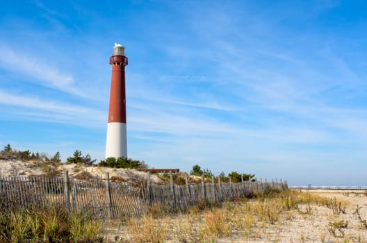 A lighthouse on the beach. There is also an old wooden fence on the beach in this landscape. This is Barnegat Lighthouse, nicknamed "Old Barney"", at Long Beach Island, New Jersey."