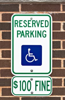 A reserved parking sign on a brick wall. There is a picture or symbol of a wheelchair on the sign along with a message for a one hundred dollar fine.