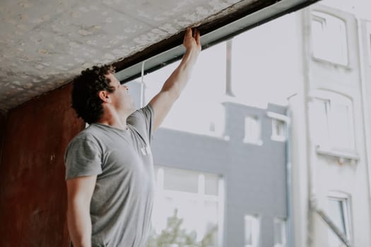A young caucasian man in a gray t-shirt with curly brown hair is repairing a window opening while standing in a half turn and cleaning the frames with his hand, preparing for the installation of a window, close-up side view. The concept of home repair, window installation, construction work, diy, lifestyle, at home.