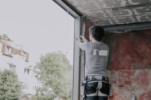 A young Caucasian man in a gray T-shirt with curly brown hair stands on a stepladder and removes gray silicone with his hand from a window opening to install the frame, close-up view from below with depth of field. The concept of installing windows, construction work, house renovation.