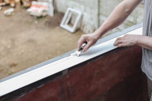 A young unrecognizable Caucasian man holds a construction knife in his hands and cleans plastic from silicone with it, preparing a window opening for window installation, close-up side view. The concept of home renovation, installation of windows, construction work.