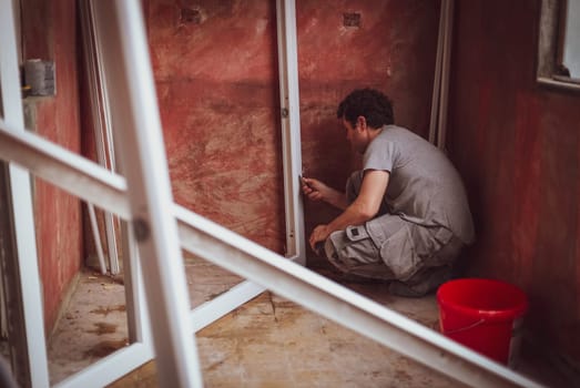 Handsome young caucasian man with curly hair sitting on the floor washes window frames with a sponge and soap against a red wall, preparing them for installation, close-up side view. The concept of home renovation, installation of windows, construction work.