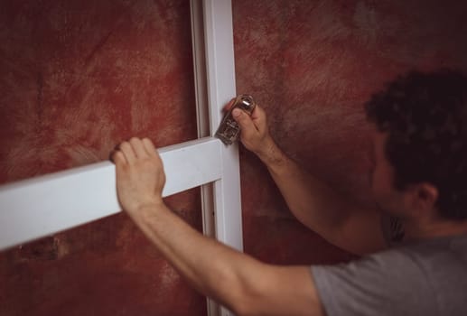 A young caucasian man with curly hair cleans the cracks on the window frame with a construction knife, preparing them for installation, close-up side view. The concept of home renovation, installation of windows, construction work.