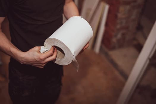 One young caucasian male builder in black uniform holds a roll of paper towels in his hands, close-up top view. The concept of home renovation, installation of windows, construction work.