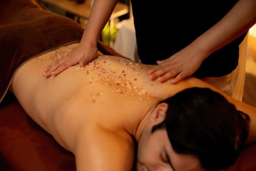 Man customer having exfoliation treatment in luxury spa salon with warmth candle light ambient. Salt scrub beauty treatment in Health spa body scrub. Quiescent