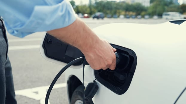 Progressive businessman install charger plug from charging station to his electric car before driving around city center. Eco friendly rechargeable car powered by sustainable and clean energy.