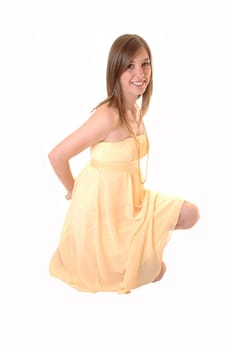 A beautiful teenager in a yellow dress kneeling on the floor in the studio holding her hands behind her back and smiling into the camera, over white.