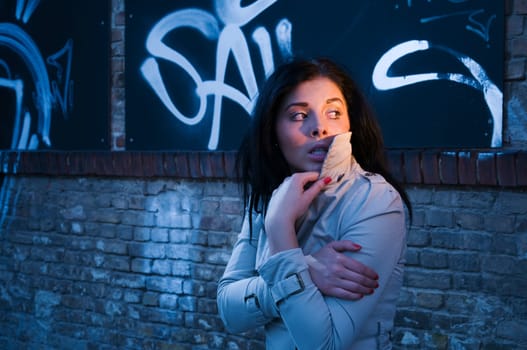 young black-haired woman in trench coat alone in the evening on a street with graffiti on a brick wall