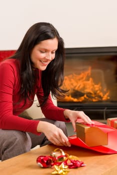 Happy woman in red wrapping Christmas present by home fireplace