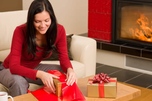 Happy woman in red wrapping Christmas present by home fireplace