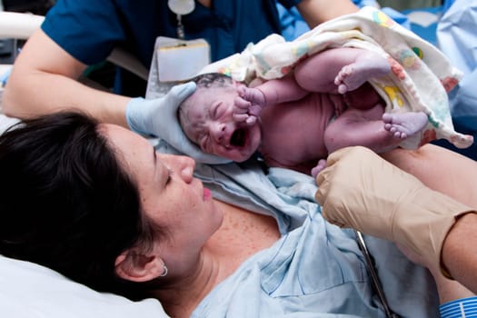 Mother in labor giving birth to baby new life in hospital. Infant covered in grease and lanugo is crying and held by nurses and doctors.