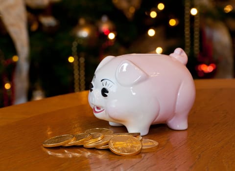 Pink piggybank on a wooden table in front of a christmas tree with gold coins