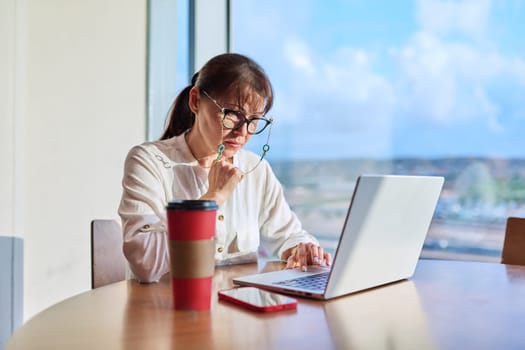 Middle-aged woman in an airport cafe working with a laptop computer, sitting at table with takeaway coffee, near panoramic window