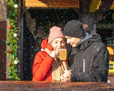 Happy student girl and boy drinking hot mulled wine and smiling at Christmas market at a snowy day. High quality photo. Photo of joyful young couple speending weekend together in winter. Portrat of two posivite amazed partners drinking a hot drink at