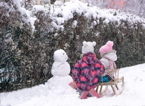 Two sisters on sled looking to snowman at snow day in park. High quality photo