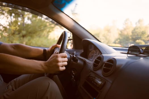 A young man travels in his car, drives along the road at sunset, holds the steering wheel with his hands, closeup view.