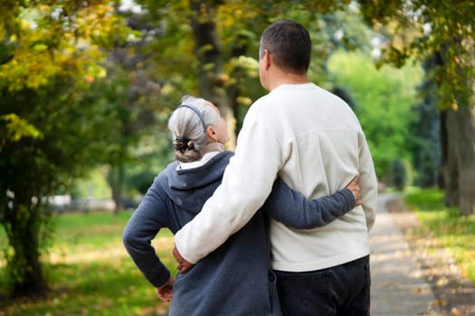 An embracing elderly couple walks in the autumn park on a sunny day, a man hugs his beloved woman and wife, people actively spend time together, develop a harmonious relationship with love.
