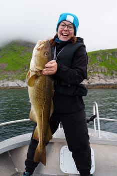 Happy young woman holding big arctic cod. Norway happy fishing. Happy fisherwoman with cod fish in hands. Guided fishing concept. Fishing tourism. Active vacation, open sea, ocean fishing.