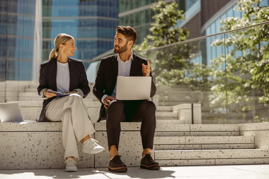 Man and woman in classic suit discussing business details and using laptop while sitting outdoors