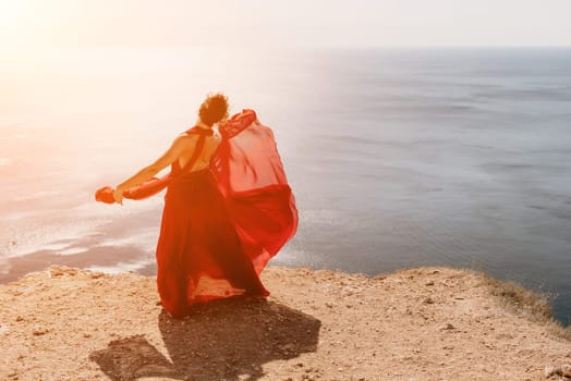 Side view a Young beautiful sensual woman in a red long dress posing on a rock high above the sea during sunrise. Girl on the nature on blue sky background. Fashion photo.