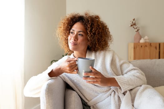 Pensive multiracial woman relaxing at home, sitting on the sofa drinking tea. Lifestyle concept.