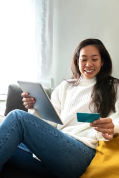 Vertical portrait of happy and smiling young Asian woman online shopping using digital tablet and credit card sitting on sofa at home. Copy space. E-commerce concept.