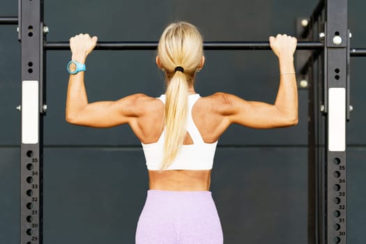 Back view of anonymous female athlete doing pulls up on horizontal bar during workout in gym