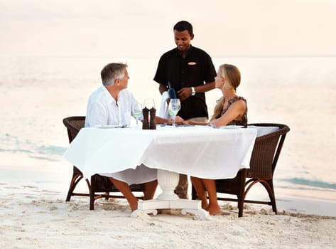 Beach, dinner and couple with sunset, love and romance with anniversary, waiter and vacation. People, romantic meal or man with woman, ocean and waves with summer holiday, travel or tropical island.