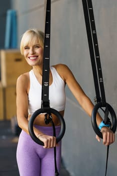 Strong female athlete in sportswear smiling while doing exercise on gymnastic rings during workout in gym
