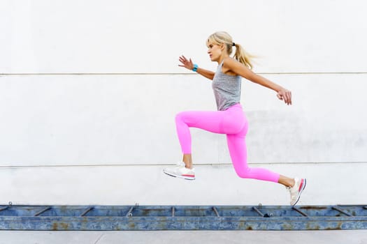 Full body side view of female athlete in pink leggings and white sneakers, looking away while jogging during running exercise on street next to shabby rusting iron ladder in daylight