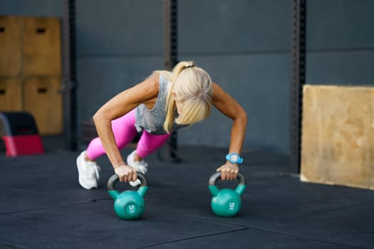 Anonymous slim sportswoman in sportswear with smartwatch looking down while doing push ups exercise with kettlebells on gym floor in daylight