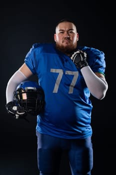 An Asian man with a red beard in a blue american football uniform holds a helmet on a black background