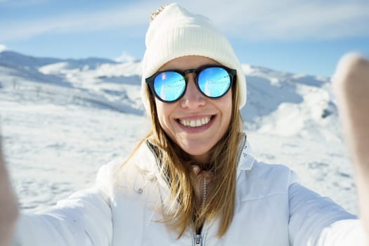 Positive young female in warm clothes beanie cap, and polarized sunglasses smiling and looking at camera while taking selfie and standing on snowy mountain slope on sunny day in winter