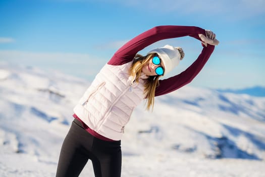 Positive young female in sportswear and sunglasses stretching arms sideway in yoga pose and looking at camera while standing on background of snowy mountains