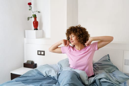 Sleepy female, with curly hair, stretching her arms on bed while getting up in early morning at home