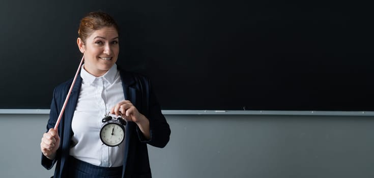 Caucasian female teacher in a pantsuit holds a pointer and a school alarm clock near the blackboard