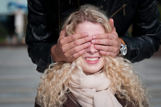 Hands of man covering eyes of young smiling blond woman - outdoor