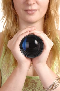 A blond young woman holding a photo lens with both hands for her chest, with reflection in the lens.