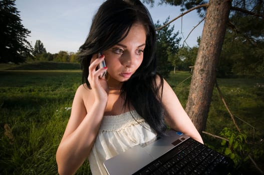 Outdoor shot of a young black-haired woman with laptop on her arm and mobile phone on her ear looking towards the screen at dusk