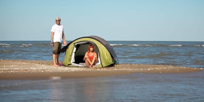happy young couple sitting on a beach next to their tent, smiling and having a good time