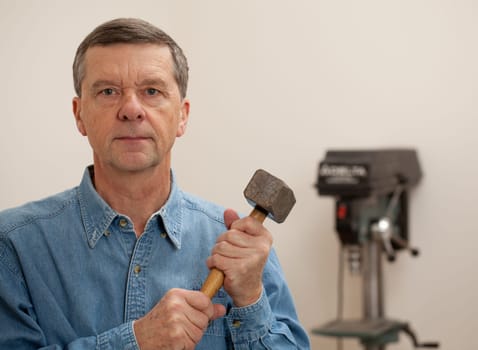 Senior male in a home workshop facing the camera and holding a large lump hammer