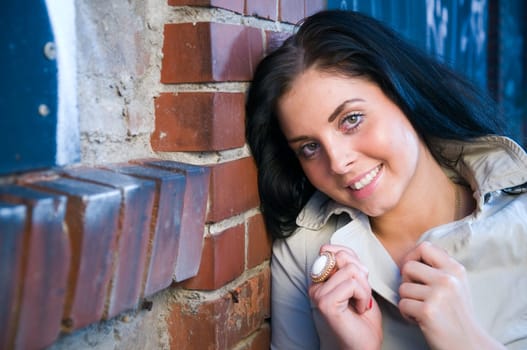 Head and shoulders portrait of a young black-haired woman with long hair on a brick wall with graffiti,hands holding up the collar of the trench coat