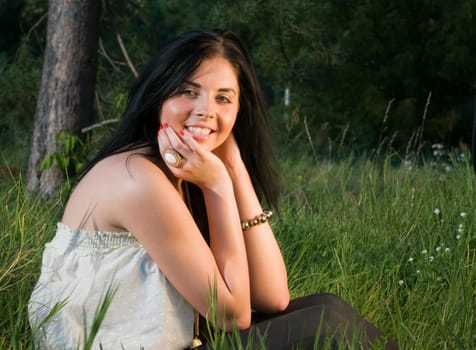 Young black-haired woman sitting in the grass resting elbows on knees and smiling at camera