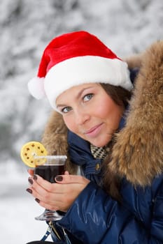 The beautiful girl in Santa&#039 s cap drinks mulled wine in winter wood.