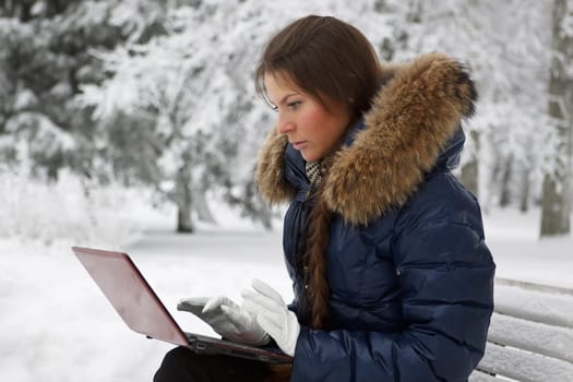 The beautiful girl with the laptop sits on a bench in winter park.