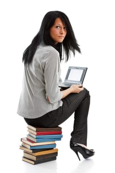The attractive student sits on a pile of books with the laptop on a white background.