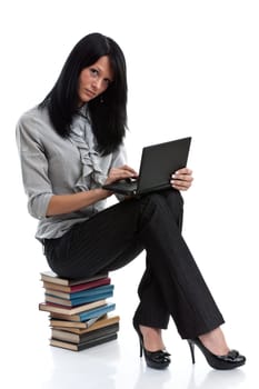 The attractive student sits on a pile of books with the laptop on a white background.