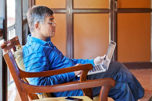 horizontal portrait of middle aged asian gentleman sat in captains chair on a a veranda with lap top computer and mobile phone, copy space and crop margins