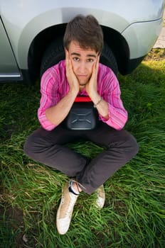 Young man sitting at the front wheel of a vehicle with a black gasoline canister and looking helplessly at the camera